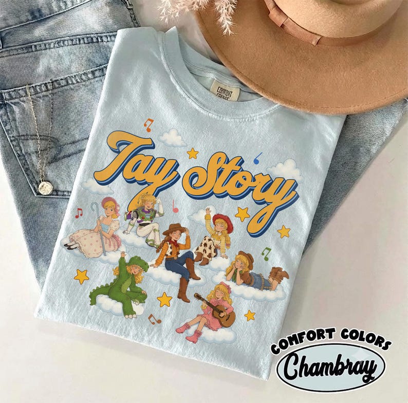 May include: A light blue t-shirt featuring the text Tay Story in yellow and blue script. The graphic includes multiple illustrated figures dressed in various costumes, including a dinosaur, a cowgirl, and a space ranger, all positioned on white clouds with yellow stars and musical notes.