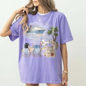 May include: A lilac-colored t-shirt featuring a beach scene with a cruise ship, palm trees, and cartoon characters. The graphic includes a sandcastle, beach chairs, and a Mickey Mouse-shaped float. The shirt has short sleeves and a relaxed fit.