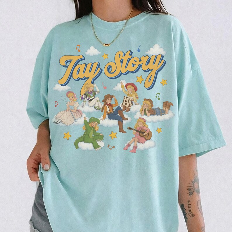 May include: A light blue short-sleeve t-shirt featuring a graphic print with the text "Toy Story" in yellow cursive font. The design includes multiple illustrated characters sitting on white clouds, surrounded by yellow stars and musical notes.