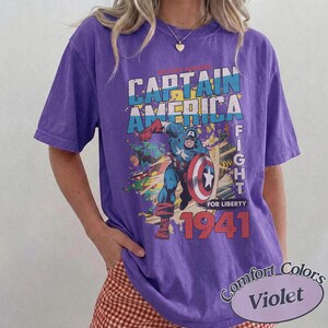 Puede incluir: Una camiseta violeta de manga corta con un estampado gr&aacute;fico de Capit&aacute;n Am&eacute;rica sosteniendo un escudo. El texto dice CAPTAIN AMERICA, FIGHT FOR LIBERTY y 1941. El dise&ntilde;o incluye colores azul, rojo y amarillo, con un estilo de c&oacute;mic desgastado.
