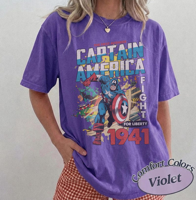 Puede incluir: Una camiseta violeta de manga corta con un estampado gr&aacute;fico de Capit&aacute;n Am&eacute;rica sosteniendo un escudo. El texto dice CAPTAIN AMERICA, FIGHT FOR LIBERTY y 1941. El dise&ntilde;o incluye colores azul, rojo y amarillo, con un estilo de c&oacute;mic desgastado.