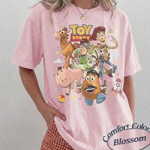 May include: A light pink t-shirt featuring a graphic print of Toy Story characters including Woody, Buzz Lightyear, Bullseye, Hamm, and Mr. Potato Head. The text Toy Story is printed in yellow and red above the characters, with small stars and a rocket.