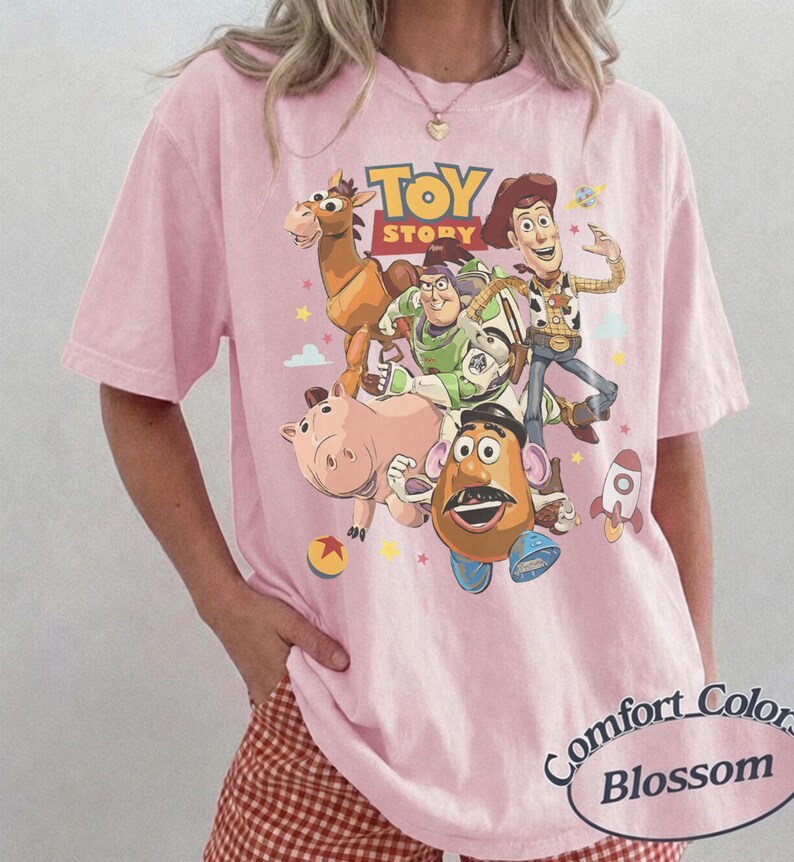 May include: A light pink t-shirt featuring a graphic print of Toy Story characters including Woody, Buzz Lightyear, Bullseye, Hamm, and Mr. Potato Head. The text Toy Story is printed in yellow and red above the characters, with small stars and a rocket.