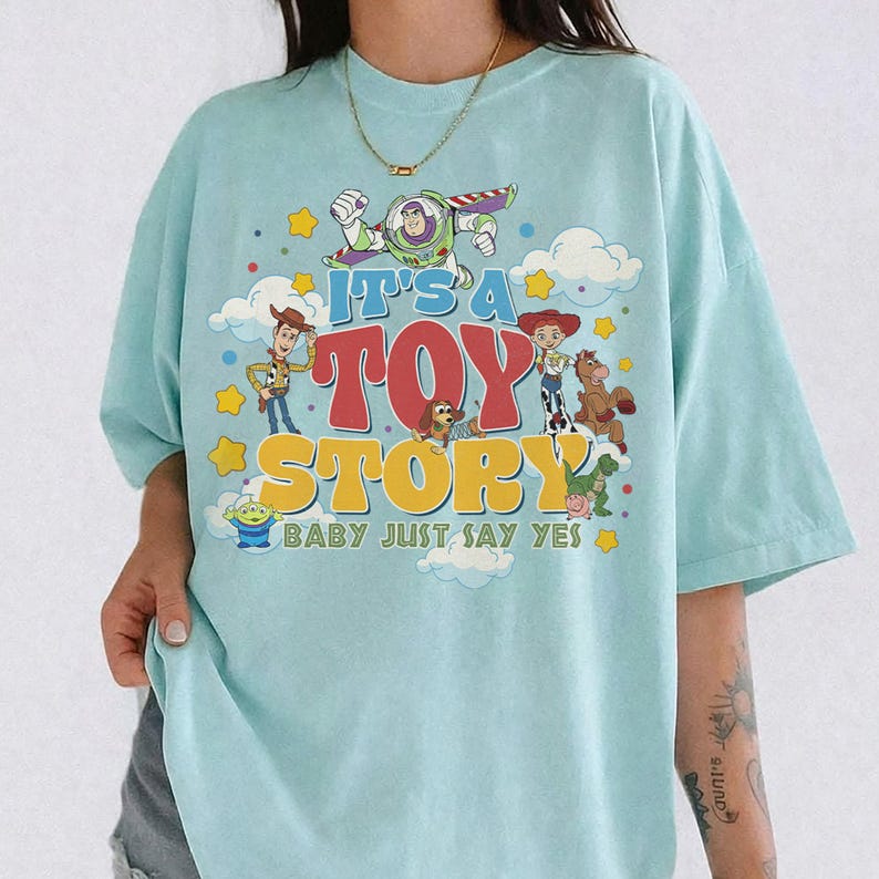 May include: A light blue t-shirt featuring a graphic print of Toy Story characters including Buzz Lightyear, Woody, Jessie, Bullseye, Slinky Dog, Rex, and an Alien. The text reads IT'S A TOY STORY BABY JUST SAY YES in blue, red, and yellow lettering.