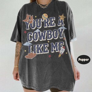 May include: A charcoal gray oversized t-shirt featuring the text YOU'RE A COWBOY LIKE ME in blue letters. The graphic includes illustrations of cowboy boots, a sheriff star, a ball, clouds, a rocket, and small stars in red, yellow, and white.