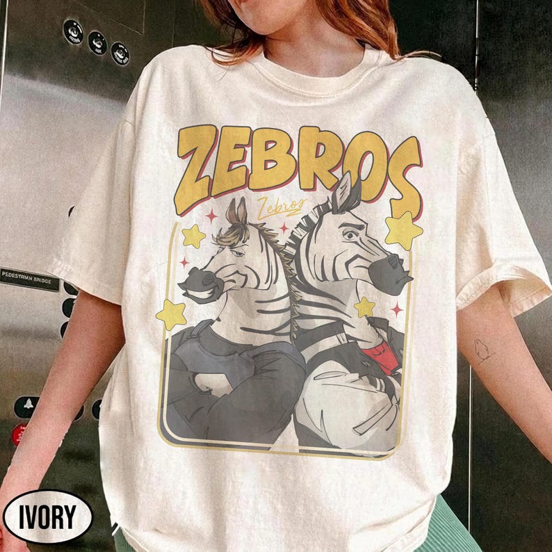 Op de afbeelding: Ivoor kleurig t-shirt met een afbeelding van twee zebra's met mensachtige trekken, omlijst door een gele rand. Het woord "ZEBROS" staat in een gele retro-lettertype boven de zebra's gedrukt. Het shirt heeft korte mouwen.