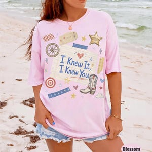 May include: A light pink short-sleeve t-shirt featuring a central graphic of a lined notepad page with the text "I Knew It, I Knew You" in blue. The design includes a cowboy boot, a sheriff star, a wagon wheel, and various small decorative elements.