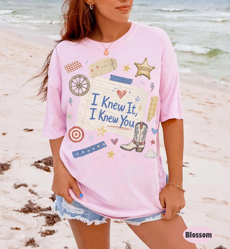 May include: A light pink short-sleeve t-shirt featuring a central graphic of a lined notepad page with the text "I Knew It, I Knew You" in blue. The design includes a cowboy boot, a sheriff star, a wagon wheel, and various small decorative elements.