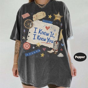 May include: A charcoal gray short-sleeve t-shirt featuring a central graphic of a lined paper scrap with the text I Knew It, I Knew You. The design includes a cowboy boot, a sheriff star, a wagon wheel, hearts, stars, and blue tape accents.