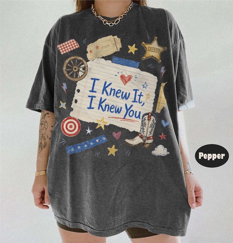 May include: A charcoal gray short-sleeve t-shirt featuring a central graphic of a lined paper scrap with the text I Knew It, I Knew You. The design includes a cowboy boot, a sheriff star, a wagon wheel, hearts, stars, and blue tape accents.