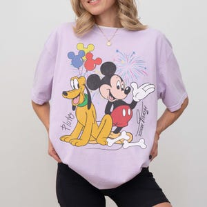 May include: A light purple t-shirt featuring a graphic of Mickey Mouse and Pluto. Mickey Mouse is wearing red shorts and Pluto is yellow. The shirt also has fireworks and balloons.