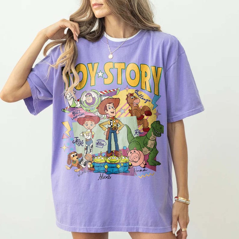 May include: A lilac t-shirt with the words "Toy Story" in yellow, featuring cartoon characters from the film. The design includes Buzz Lightyear, Woody, Jessie, and other characters. The shirt has short sleeves and a relaxed fit.