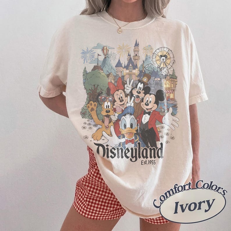 Puede incluir: Camiseta de color marfil con un gr&aacute;fico de Disneyland que presenta a Mickey Mouse, Minnie Mouse, Donald Duck, Goofy y Pluto. El gr&aacute;fico incluye un castillo, fuegos artificiales y una noria. El texto "Disneyland Est. 1955" est&aacute; debajo de los personajes. La camiseta est&aacute; etiquetada como "Comfort Colors Ivory".