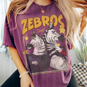 Op de afbeelding: Paars T-shirt met het woord "ZEBROS" in gele letters boven een illustratie van twee zebra's. De zebra's dragen kleding en worden omlijst door een gouden rand met gele sterren. Het shirt heeft een vintage, vervaagde uitstraling.