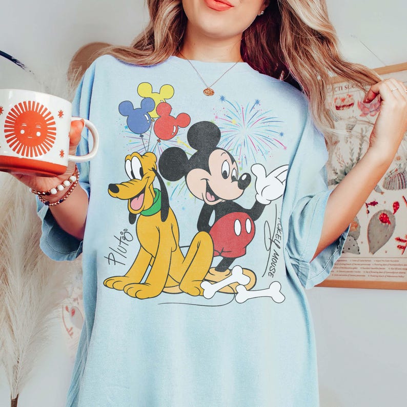 May include: Light blue t-shirt featuring Mickey Mouse and Pluto with colorful balloons and fireworks. Mickey Mouse is wearing red shorts and white gloves. Pluto is yellow. The shirt has the text "Mickey Mouse" and "Pluto".