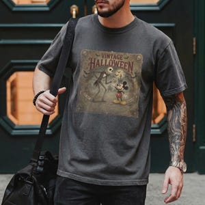 May include: A charcoal gray short-sleeve t-shirt featuring a graphic print with the text "VINTAGE HALLOWEEN". The design depicts Jack Skellington and Mickey Mouse standing in front of a castle, surrounded by bats and skulls in a muted color palette.
