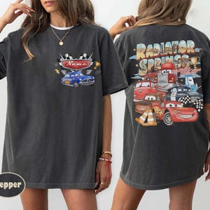 May include: Dark grey t-shirt with a front graphic of a blue car and the word "Name". The back of the shirt has a colourful graphic of cartoon cars and the text "Radiator Springs".