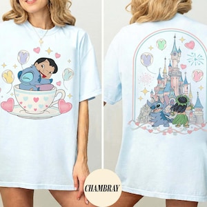 Puede incluir: Camiseta azul claro con un dise&ntilde;o de dibujos animados de un personaje en una taza de t&eacute; con globos y corazones. La parte posterior de la camiseta muestra un castillo, fuegos artificiales y personajes. La palabra "CHAMBRAY" est&aacute; impresa en la parte inferior derecha.