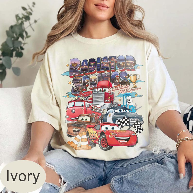 May include: Ivory t-shirt with a colourful graphic of Cars movie characters, including Lightning McQueen and the words "Radiator Springs." The design includes a trophy and chequered flags.