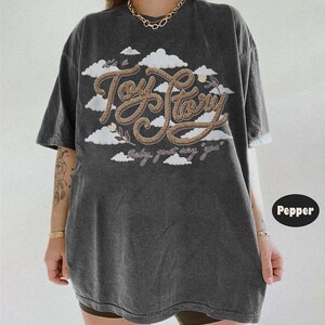 Pu&ograve; includere: Una t-shirt oversize grigio antracite con una grafica di nuvole bianche e il testo "it's a Toy Story" scritto in un font marrone stile corda, con il testo "baby, just say 'yea'" stampato sotto in un carattere corsivo pi&ugrave; piccolo.