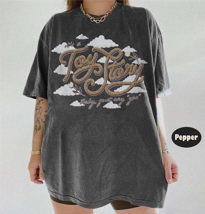 Pu&ograve; includere: Una t-shirt oversize grigio antracite con una grafica di nuvole bianche e il testo "it's a Toy Story" scritto in un font marrone stile corda, con il testo "baby, just say 'yea'" stampato sotto in un carattere corsivo pi&ugrave; piccolo.