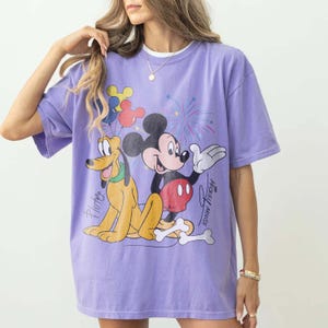 May include: A lilac-colored t-shirt featuring a graphic of Mickey Mouse and Pluto with fireworks and balloons. Mickey Mouse is in a red and black outfit, and Pluto is yellow. The words "Mickey Mouse" and "Pluto" are written on the shirt.