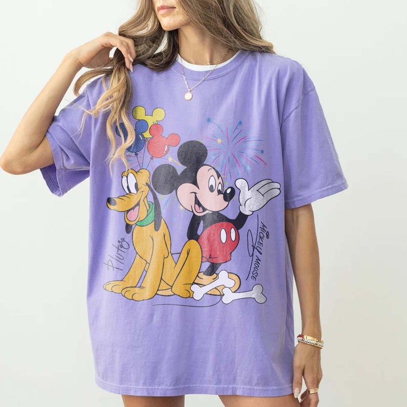 May include: A lilac-colored t-shirt featuring a graphic of Mickey Mouse and Pluto with fireworks and balloons. Mickey Mouse is in a red and black outfit, and Pluto is yellow. The words "Mickey Mouse" and "Pluto" are written on the shirt.
