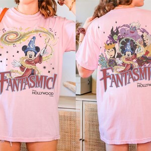 May include: Pink t-shirt featuring Mickey Mouse as a sorcerer with the text "Fantasmic!" in a stylized font. The back of the shirt displays various Disney villains. The shirt is a light pink color.