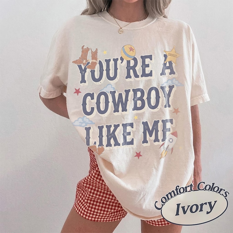 May include: An ivory short-sleeve t-shirt featuring the blue text "YOU'RE A COWBOY LIKE ME" printed on the front. The design includes illustrations of brown cowboy boots, a yellow ball, a sheriff star, blue clouds, red stars, and a rocket ship.