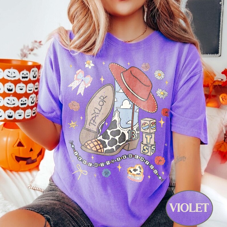 Puede incluir: Una camiseta morada con un estampado gr&aacute;fico de una bota vaquera con manchas de vaca, un sombrero rojo, un lazo y bloques con el texto "13" y "TS". El dise&ntilde;o incluye el texto "TAYLOR" en la suela de la bota y "I KNEW IT I KNEW YOU".