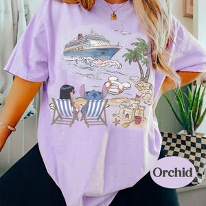 May include: A light purple t-shirt featuring a cruise ship on the ocean, palm trees, and a beach scene with cartoon characters. The shirt has a relaxed fit and the word "Orchid" printed on the bottom right.