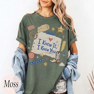 May include: A moss green t-shirt featuring a central graphic of a torn piece of notebook paper with the text I Knew It, I Knew You. The design includes a cowboy boot, a sheriff star, a wagon wheel, a target, and various small stars and hearts.