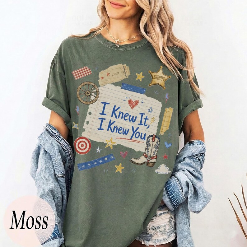 May include: A moss green t-shirt featuring a central graphic of a torn piece of notebook paper with the text I Knew It, I Knew You. The design includes a cowboy boot, a sheriff star, a wagon wheel, a target, and various small stars and hearts.