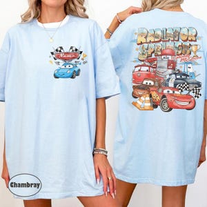 May include: Pale blue t-shirt with a cartoon car design on the front and a "Radiator Springs" graphic on the back. The front design includes a blue car, checkered flags, and the word "Name". The back design features various cartoon cars.