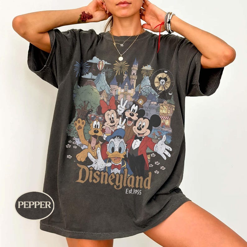 Puede incluir: Camiseta gris oscuro con un gr&aacute;fico colorido de personajes de Disney, incluyendo Mickey Mouse, Minnie Mouse, Donald Duck y Goofy, frente a un castillo y fuegos artificiales. La palabra "Disneyland" y "Est.1955" est&aacute;n impresas en la camiseta.