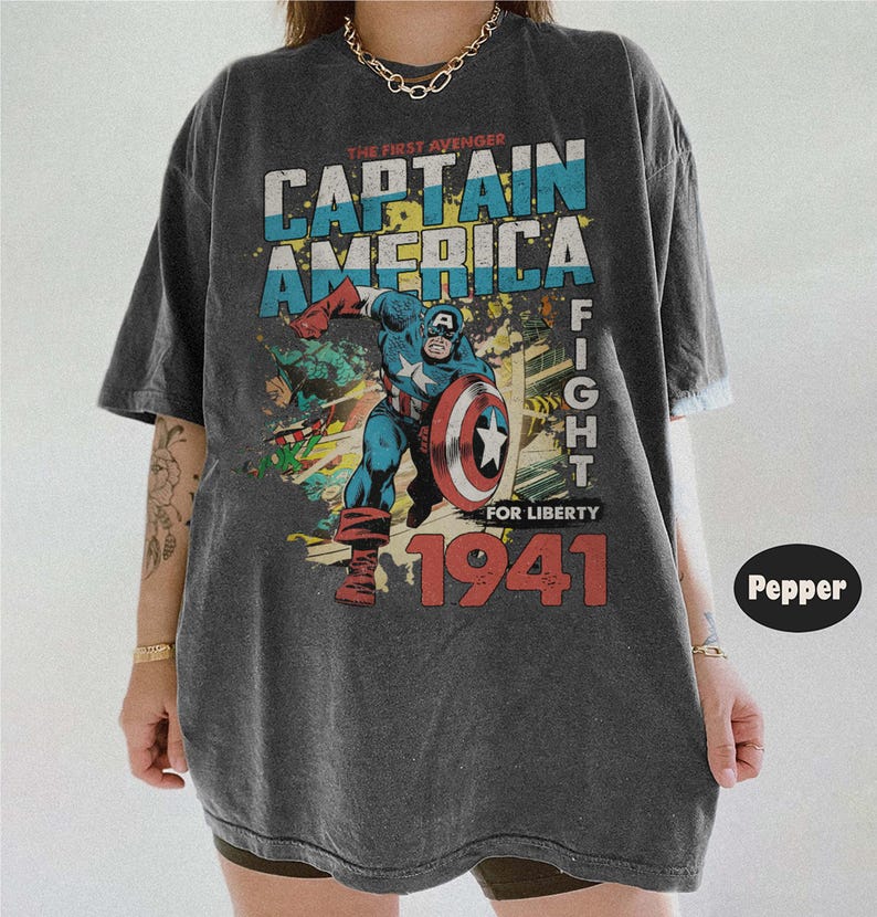 Puede incluir: Una camiseta gris carb&oacute;n de corte oversize con un gr&aacute;fico de estilo vintage de Capit&aacute;n Am&eacute;rica en una pose de combate din&aacute;mica. El estampado incluye el texto THE FIRST AVENGER, CAPTAIN AMERICA, FIGHT FOR LIBERTY y 1941 en letras grandes.