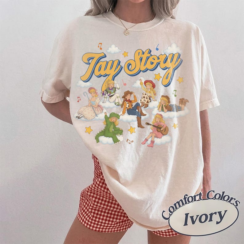 May include: An ivory short-sleeve t-shirt featuring a graphic print with the text Tay Story in yellow cursive letters. The design includes multiple illustrated figures dressed in various outfits sitting on white clouds with yellow stars and musical notes.