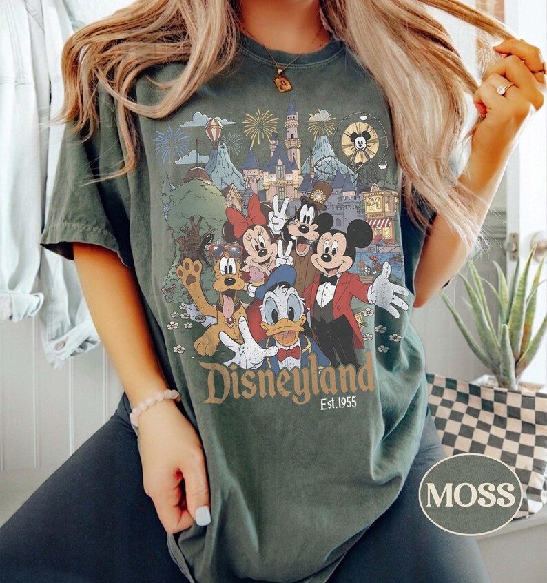 Puede incluir: Camiseta verde musgo con un gr&aacute;fico de Disneyland. El dise&ntilde;o incluye a Mickey Mouse, Minnie Mouse, Donald Duck y Goofy, junto con un castillo, fuegos artificiales y el texto "Disneyland Est. 1955".