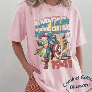 Puede incluir: Una camiseta de manga corta de color rosa claro con un estampado gr&aacute;fico de Capit&aacute;n Am&eacute;rica en una pose din&aacute;mica con un escudo. El texto dice THE FIRST AVENGER CAPTAIN AMERICA FIGHT FOR LIBERTY 1941. Una etiqueta ovalada dice Comfort Colors Blossom.