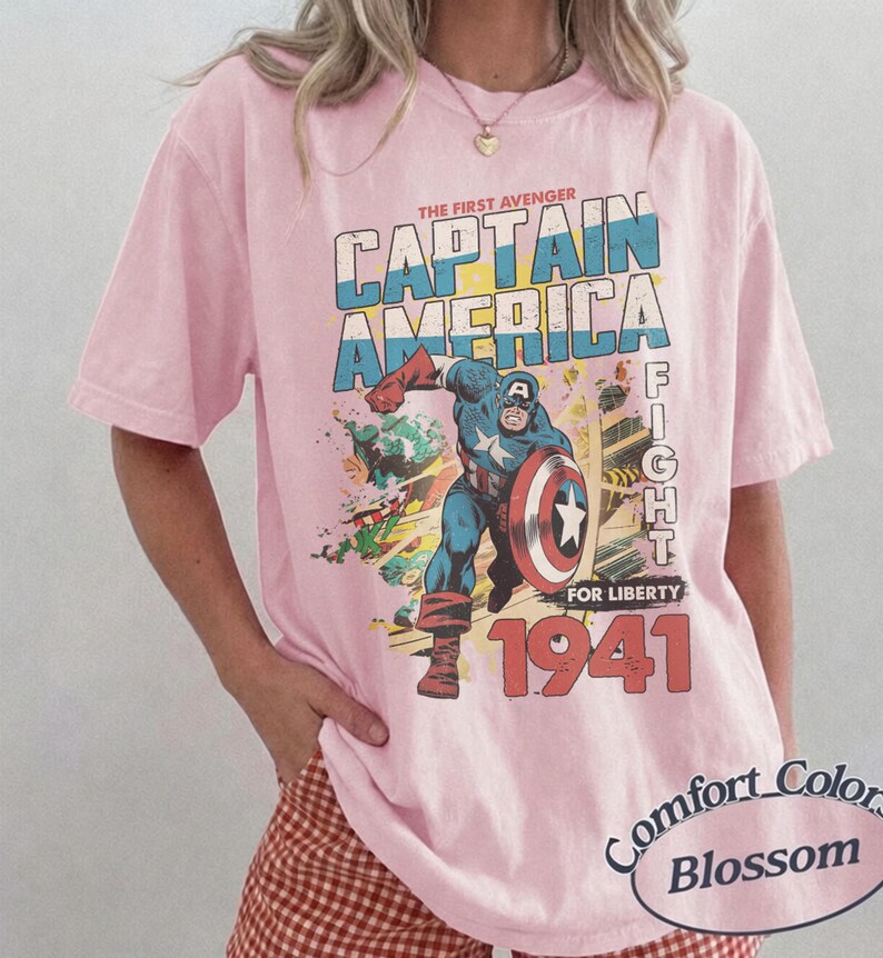 Puede incluir: Una camiseta de manga corta de color rosa claro con un estampado gr&aacute;fico de Capit&aacute;n Am&eacute;rica en una pose din&aacute;mica con un escudo. El texto dice THE FIRST AVENGER CAPTAIN AMERICA FIGHT FOR LIBERTY 1941. Una etiqueta ovalada dice Comfort Colors Blossom.