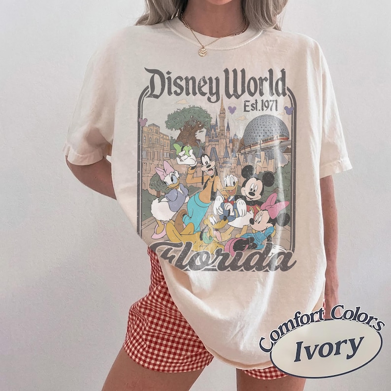 May include: Ivory-colored t-shirt featuring a Disney World graphic with Mickey Mouse, Minnie Mouse, Donald Duck, Daisy Duck, and Goofy. The graphic includes the text "Disney World Est. 1971 Florida". The shirt is a Comfort Colors brand.