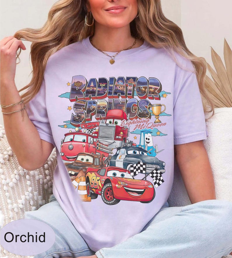 May include: Orchid-coloured t-shirt featuring a graphic of characters and the text "Radiator Springs Lightning McQueen". The design includes various cartoon cars, a trophy, and checkered flags. The shirt is a light purple colour.