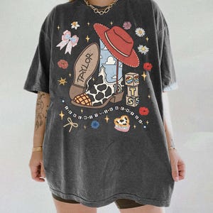 Puede incluir: Una camiseta gris carb&oacute;n de corte oversize con un estampado gr&aacute;fico de una bota vaquera con manchas de vaca, un sombrero vaquero rojo y bloques con las letras T S. El dise&ntilde;o incluye el texto TAYLOR, 13 e I KNEW IT, I KNEW YOU.