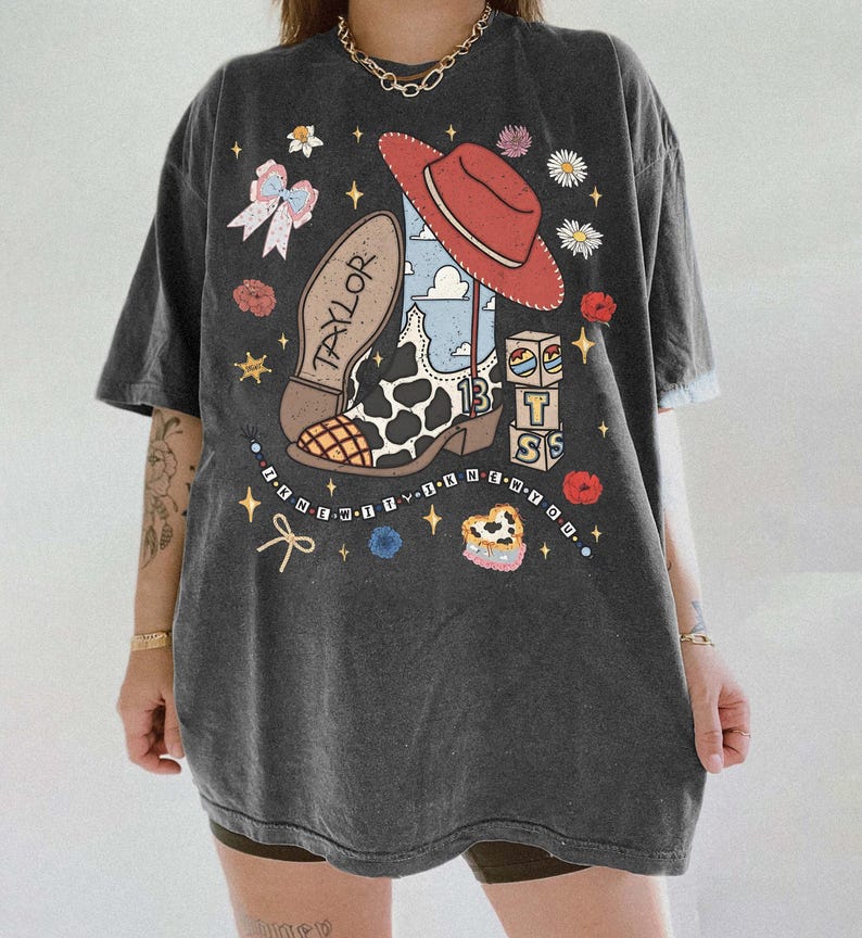 Puede incluir: Una camiseta gris carb&oacute;n de corte oversize con un estampado gr&aacute;fico de una bota vaquera con manchas de vaca, un sombrero vaquero rojo y bloques con las letras T S. El dise&ntilde;o incluye el texto TAYLOR, 13 e I KNEW IT, I KNEW YOU.
