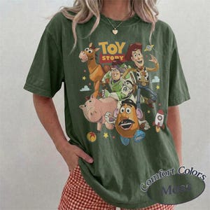 May include: An olive green short-sleeve t-shirt featuring a graphic print of Toy Story characters including Woody, Buzz Lightyear, Bullseye, Hamm, and Mr. Potato Head. The text TOY STORY is printed in yellow and red above the characters.