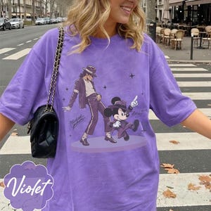 Pode incluir: Uma t-shirt roxa de manga curta com uma estampa gr&aacute;fica de uma pessoa de fedora e fato a dan&ccedil;ar ao lado de um rato de desenho animado com fedora e fato a condizer. A t-shirt inclui uma mala preta com al&ccedil;a de corrente dourada.
