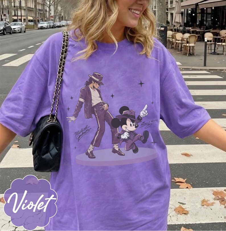 Pode incluir: Uma t-shirt roxa de manga curta com uma estampa gr&aacute;fica de uma pessoa de fedora e fato a dan&ccedil;ar ao lado de um rato de desenho animado com fedora e fato a condizer. A t-shirt inclui uma mala preta com al&ccedil;a de corrente dourada.