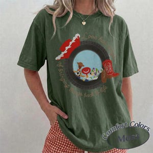May include: An olive green short-sleeve t-shirt featuring a graphic of a black tire framing a scene with cartoon characters. The design includes a red cowboy hat, a red boot, and the text Love has a way of bringing things back to life.