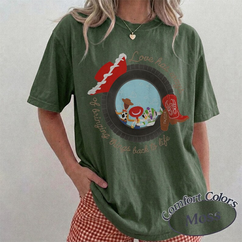 May include: An olive green short-sleeve t-shirt featuring a graphic of a black tire framing a scene with cartoon characters. The design includes a red cowboy hat, a red boot, and the text Love has a way of bringing things back to life.