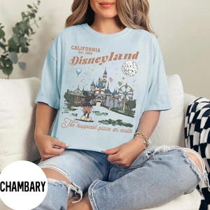 Puede incluir: Camiseta azul claro con un gr&aacute;fico de Disneyland. El dise&ntilde;o incluye el texto "California Est. 1955 Disneyland" y una ilustraci&oacute;n de Mickey Mouse, un castillo y una noria. Tambi&eacute;n se incluye la frase "The happiest place on earth".